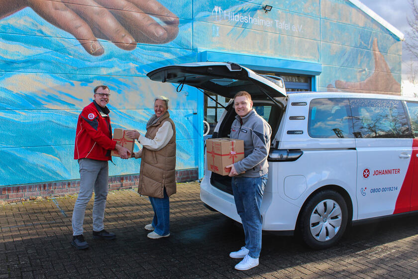 Zwei Johanniter mit Paketen neben einem Johanniter-Fahrzeug. Ein Johanniter überreicht einer Frau sein Paket. Im Hintergrund ein blaues Gebäude mit der Aufschrift "Hildesheimer Tafel"