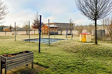 Grüne Außenfläche mit eingezäuntem Spielbereich, Bäumen, Sandkasten und einem kleinen bunten Spielhäuschen.