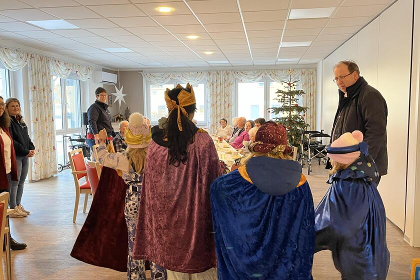 Sternsinger mit Pfarrer im großen Aufenthaltsraum der Tagespflege, wo die Gäste am runden Tisch sitzen