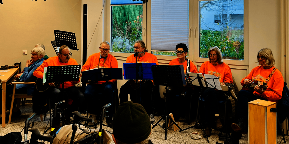 Eine Band mit mehreren Mitgliedern in orangefarbenen T-Shirts spielt gemeinsam auf Ukulelen und anderen Instrumenten in einem festlich geschmückten Raum; vor ihnen sitzen Zuschauende, einige mit Rollator.