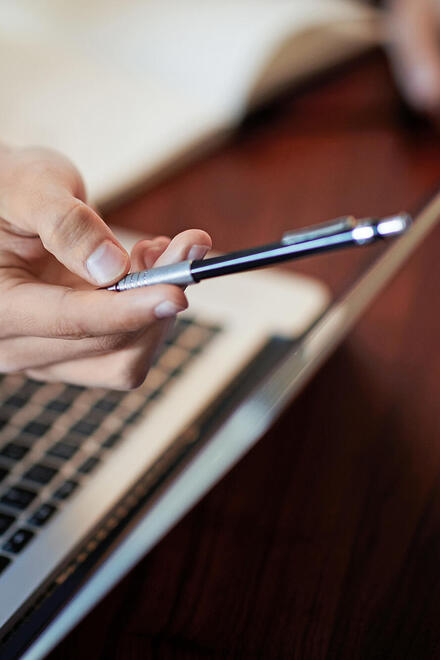 Zu sehen ist ein Laptop, davor eine Hand, die einen Stift hält