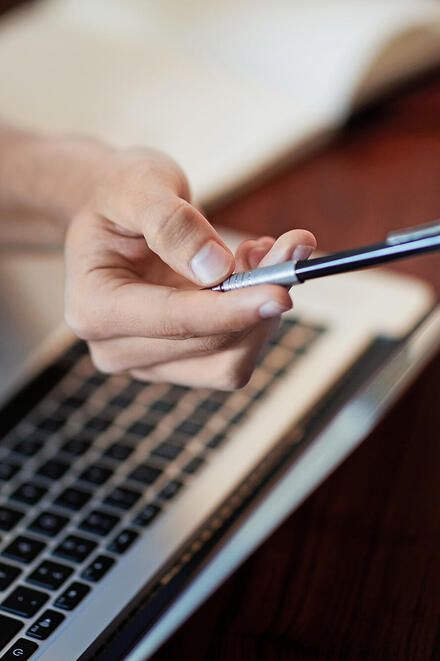 Zu sehen ist ein Laptop, davor eine Hand, die einen Stift hält