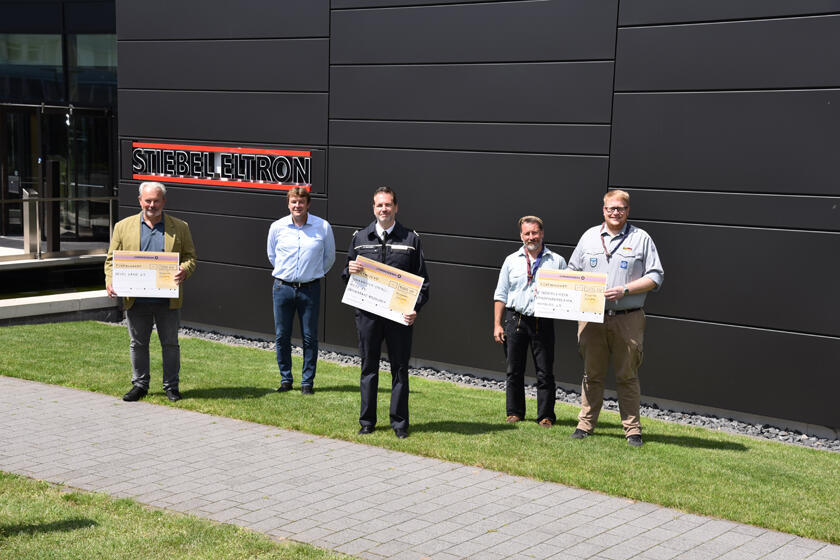 Vor dem Gelände von Stiebel Eltron stehen die Empfänger der Spenden und halten einen großen Scheck in der Hand, auf dem die jeweilige Spendensumme steht.