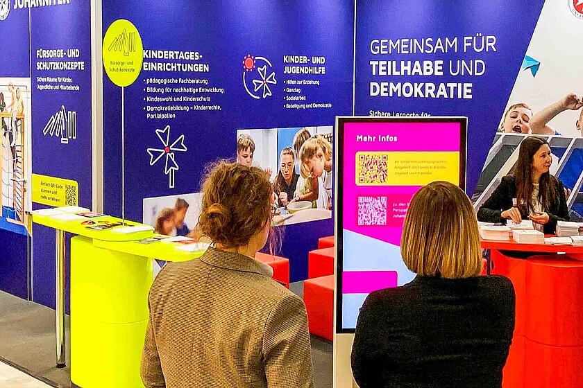 Zwei Frauen shcauen sich den farbenfroh gestalteten Stand der Johanniter bei der didacta an