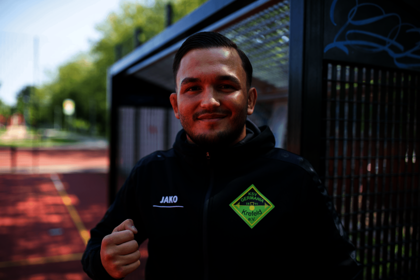 Ein Mann in schwarzer Jacke steht an einer Sportbank und lächelt