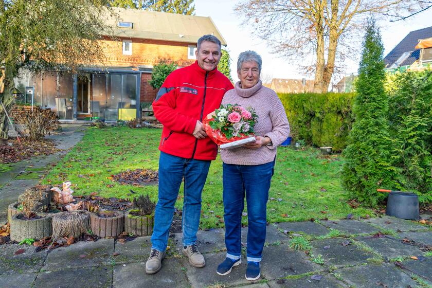 Ein Mitarbeiter der Johanniter und eine Seniorin stehen in einem Garten und lächeln in die Kamera.