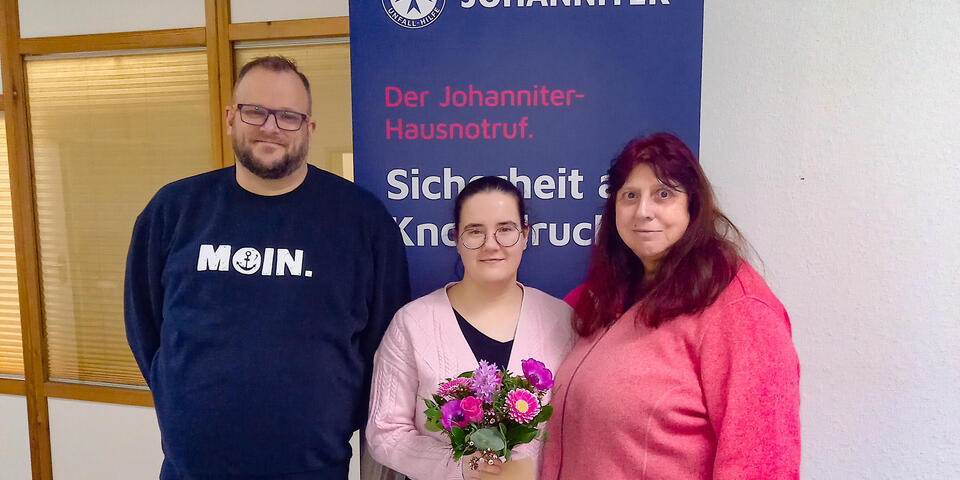 Ein Mann, eine junge Frau mit Blumenstrauß und eine weitere Frau vor einem Hausnotruf-Roll-Up