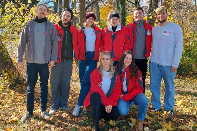 Mehrere Leute stehen in einem Wald und lächeln in die Kamera. Am Boden liegen bunte Blätter.