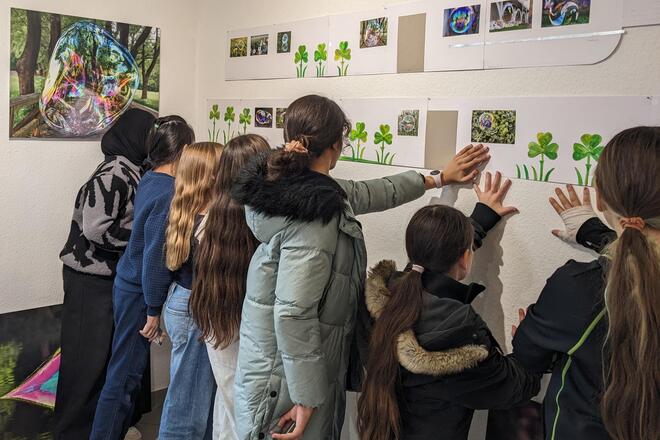 Mehrere Kinder und Jugendliche betrachten gemeinsam eine Wand mit künstlerisch gestalteten Bildern und Fotomotiven von Seifenblasen in einer Ausstellung. Einige Kinder berühren die Ausstellungsstücke, andere schauen neugierig und interessiert auf die bunten Seifenblasenmotive.