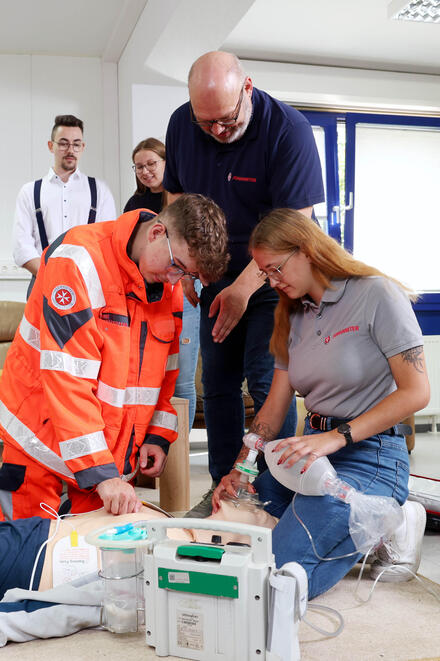 Eine Gruppe junger Menschen kniend an einer Übungspuppe. Ein Mann in Johanniter-Kleidung beugt sich zu ihnen. Andere schauen zu.