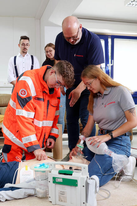 Eine Gruppe junger Menschen kniend an einer Übungspuppe. Ein Mann in Johanniter-Kleidung beugt sich zu ihnen. Andere schauen zu.