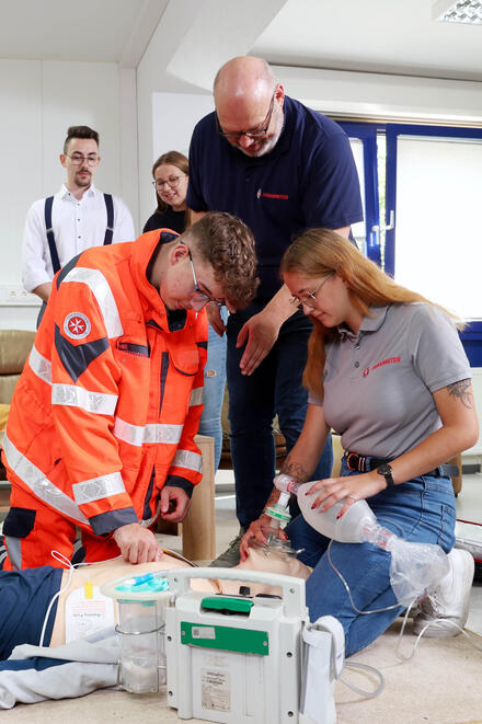 Eine Gruppe junger Menschen kniend an einer Übungspuppe. Ein Mann in Johanniter-Kleidung beugt sich zu ihnen. Andere schauen zu.