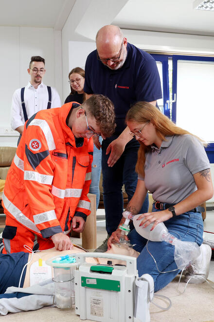 Eine Gruppe junger Menschen kniend an einer Übungspuppe. Ein Mann in Johanniter-Kleidung beugt sich zu ihnen. Andere schauen zu.