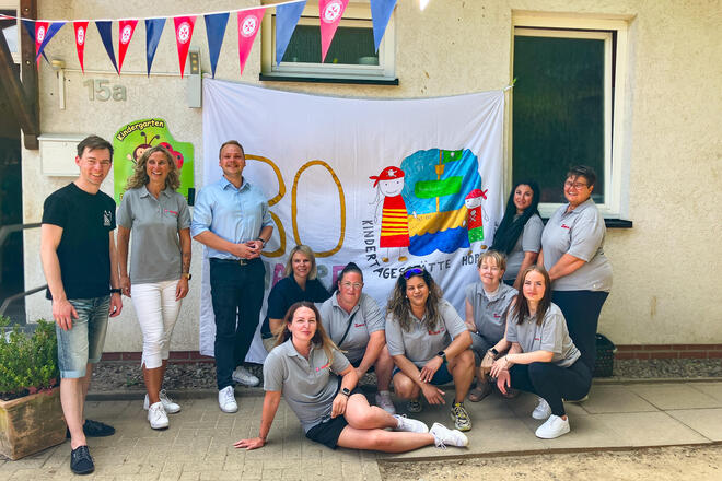 Eine Gruppe von Menschen stehen oder sitzen vor einer gelben Hauswand. Am Haus ein weißes Laken auf dem steht "30 Jahre"