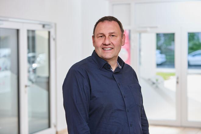 Ein Mann mit dunkelbraunen Haaren und einem dunkelblauem Hemd