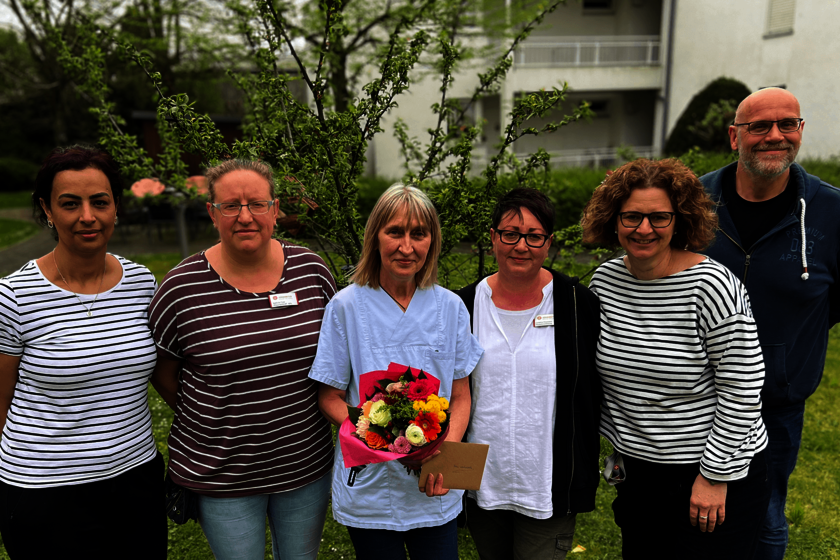 Eine Gruppe von Menschen steht nebeneinander im Grünen. Eine Frau hält einen Blumenstrauß in den Händen.