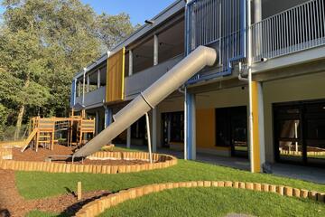 Die Außenansicht der Kita mit Außenrutsche und begrünter Außenanlage mit Klettergerüst.