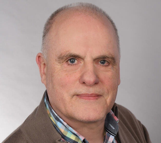 Ein Mann mit hoher Stirn und graumelierten Haaren, einem karierten Hemd und einer braunen Strickjacke