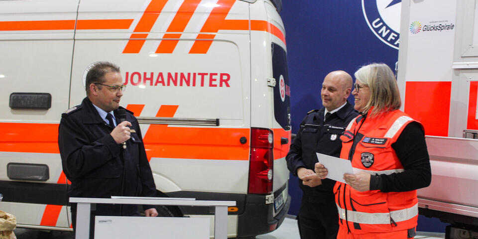 Ein Mann mit Mikro steht an einem Rednerpult und spricht zu einer Frau und einem Mann