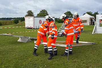 Vier Einsatzkräfte tragen eine Trage gemeinsam durch einen Hindernisparcours.