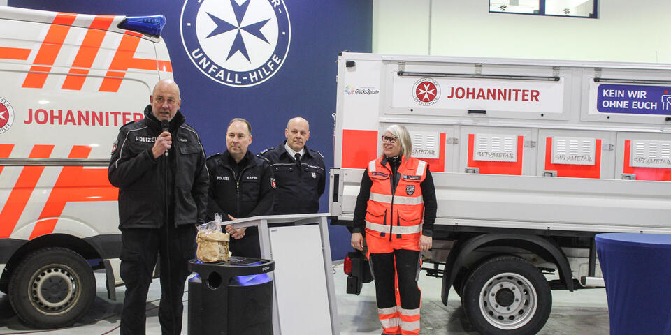 Zwei Männer, einer mit Mikro an einem Rednerpult und  einer spricht zu einer Frau und einem Mann