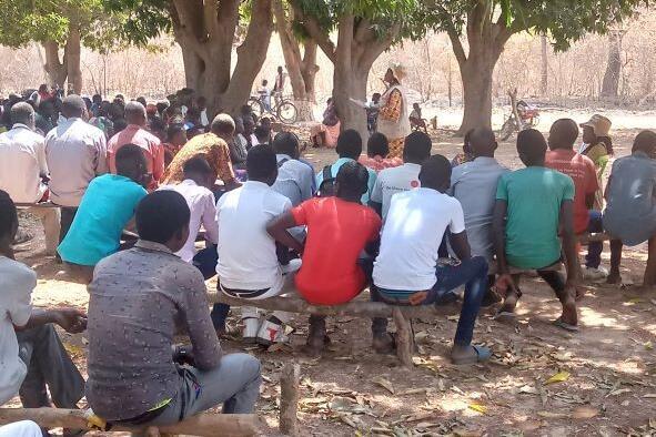 Eine Gruppe junger Männer im Vordergrund, eine Rednerin mit Johanniter Westeim Hintergrund