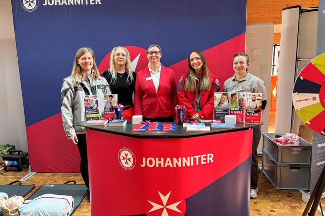 Vier Frauen in Johanniter-Kleidung an einem Messestand. Links liegt eine Reanimationspuppe auf einer Matte. Rechts ist ein Glücksrad zu sehen. 