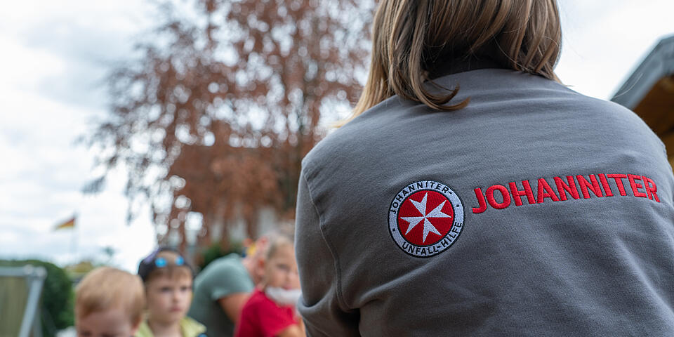 Kinder sitzen am Tisch und Johanniterin zeigt wie die T-Shirts gebatikt werden.