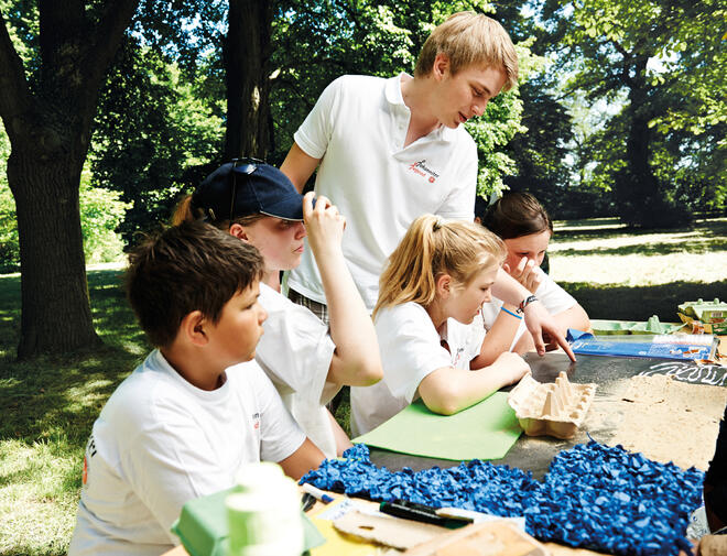 Kinder sitzen an einem Tisch ein junger Mann steht daneben und zeigt ihnen etwas