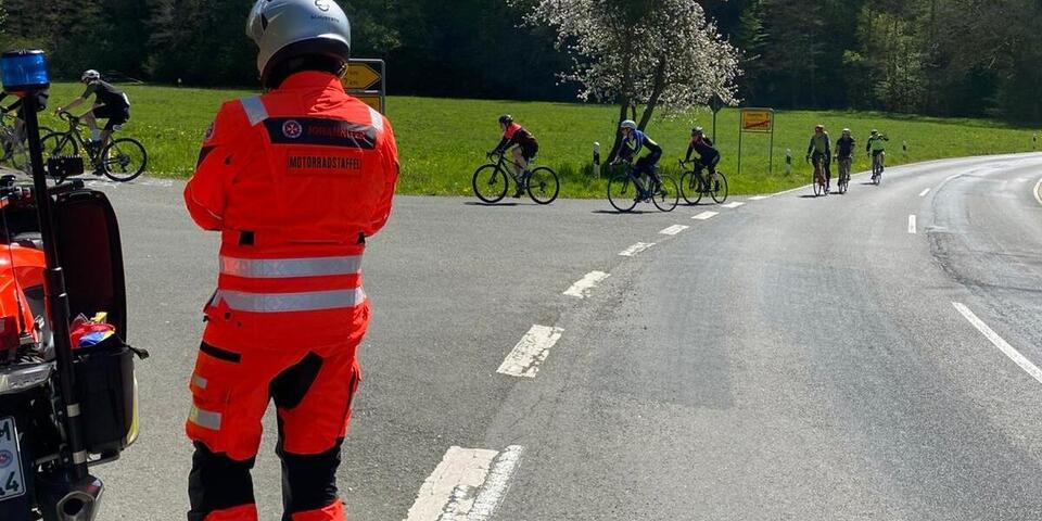 Ein Mann in Motorradbekleidung und mit Helm steht neben seinem Motorrad auf einer Landstraße. Etwas entfernt fahren Radsportler.