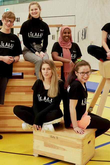 Acht junge Ferienbetreuerinnen und -betreuer posieren fröhlich in einer Turnhalle. Sie sind in sportlicher Kleidung und tragen alle einheitliche "Lilalu"-T-Shirts
