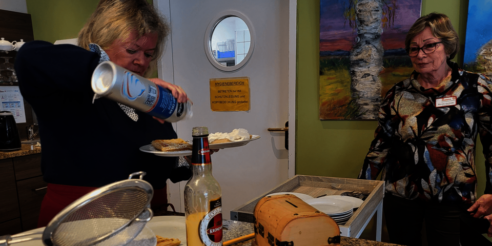 Ein Frau sprüht Sahne auf einen Kuchenteller, eine andere Frau sieht zu.