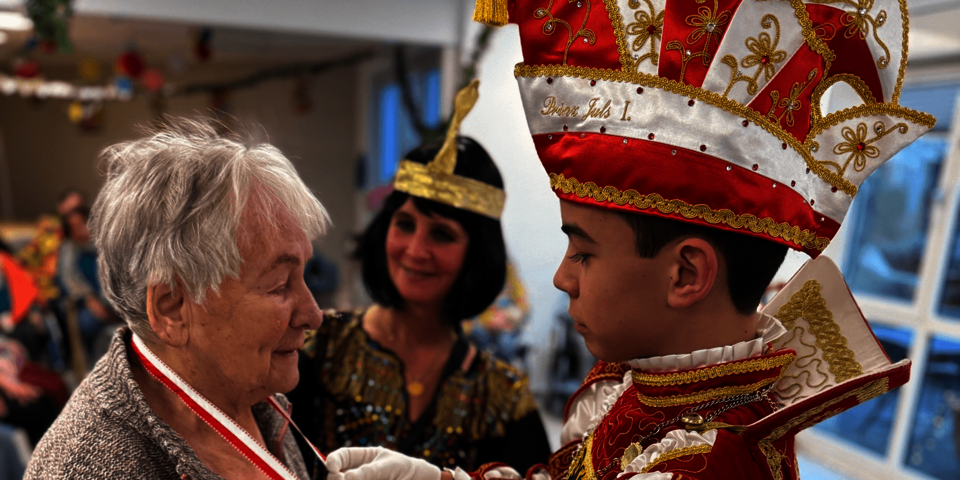 Junger Karnevalsprinz zeichnet eine ältere Dame mit einer Medaille aus, Zuschauer im Hintergrund.